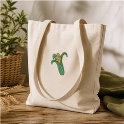 Embroidered corn cob on a light beige tote bag, surrounded by green plant leaves, evokes a fresh summer vibe.