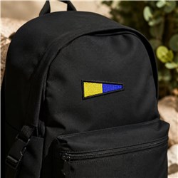 Modern black backpack with geometric yellow and blue triangles on the front pocket, set against soft greenery.