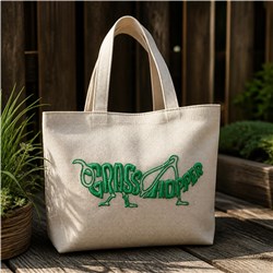 Vibrant green grasshopper graphic embroidered on a beige tote bag among natural wood and lush greenery.