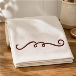Elegant folded towel with cream fabric and delicate brown scroll embroidery on a warm wooden surface.