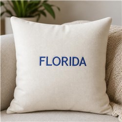 Cream pillow with bold blue 'FLORIDA' embroidery, set on a cozy sofa with subtle greenery.