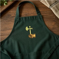 Axe hand gripping an axe embroidered on a dark green apron, with bright colors against a rustic wooden background.