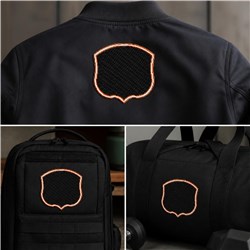 Blank shield emblem with orange stitching on black fabric, featured on a jacket, backpack, and duffel bag.