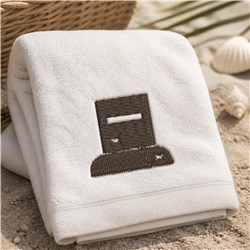 White towel with minimalistic brown embroidered design, neatly folded on sandy surface with seashells and greenery.
