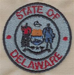 Delaware state emblem with blue circle, red lettering, two historical figures, and the state shield in the center.