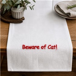 Table runner with playful text embroidery 'Beware of Cat!' in bold red on white fabric, set on a wooden table.