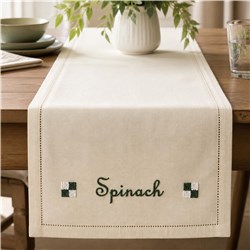 Table runner with 'Spinach' in dark green embroidery, set on a wooden table, surrounded by a vase of greenery.