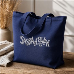 Navy blue tote bag with bold white 'SASKATCHEWAN' embroidery, displayed on wood with natural lighting.