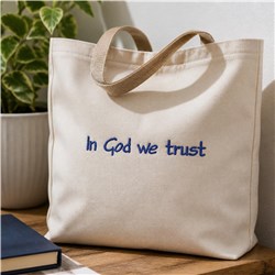 Beige tote bag with blue embroidered text 'In God we trust', simple modern design on wooden surface.