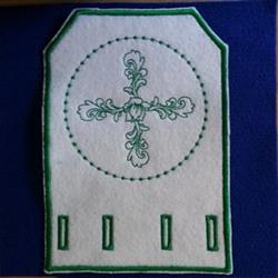 Embroidered decorative patch featuring an intricate cross design in green thread on white felt, with elegant swirls.